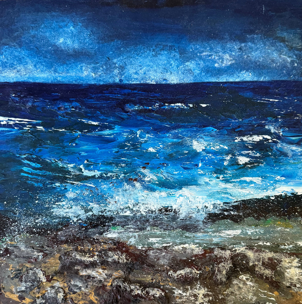 A square framed painting of a dramatic blue ocean scene with white waves crashing onto a rocky shore, displayed on a rustic wooden surface.