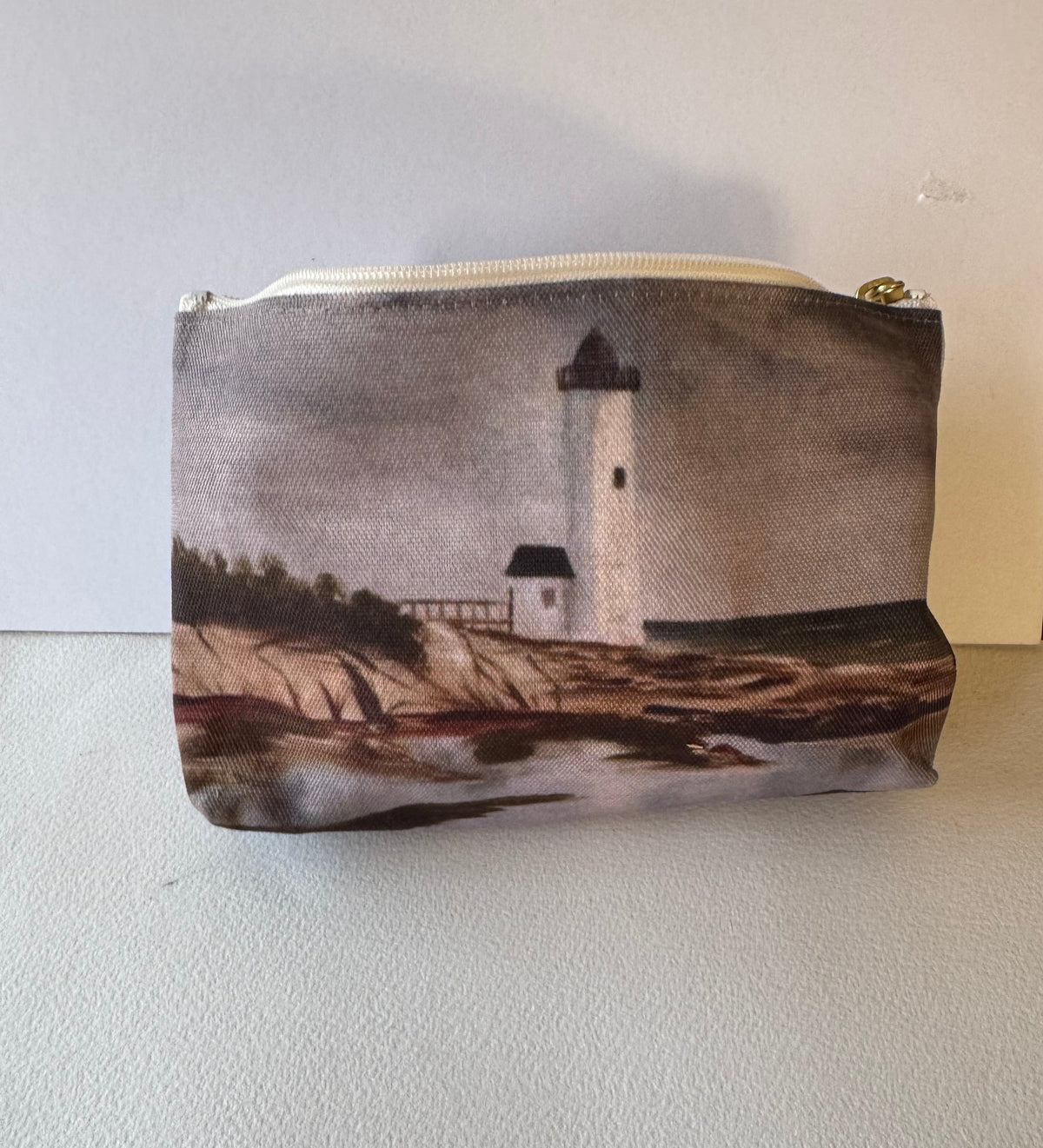 A zippered pouch featuring a printed scenic image of a lighthouse by the water, shown on a light-colored textured surface.
